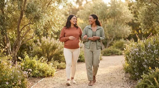A pregnant mom and her birth-care professional walking arm in arm along a sunlit garden path.